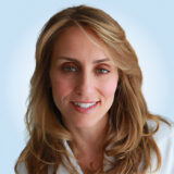 A woman with long, wavy, light brown hair and a gentle smile, wearing a white top, against a light blue background.