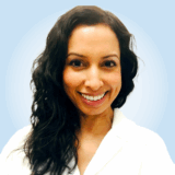 A smiling woman with long, wavy black hair is shown against a soft blue background. She wears a white coat and radiates warmth and professionalism, embodying a friendly atmosphere.