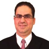 A man wearing glasses and a suit with a tie smiles confidently at the camera. He has short, dark hair and appears professional, suitable for a business or formal setting.