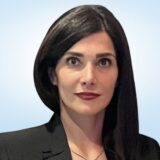 A woman with long black hair and a serious expression is wearing a black blazer. The background is a soft gradient.