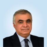 A man with short gray hair and a neutral expression is wearing a black suit and striped tie against a light blue background.