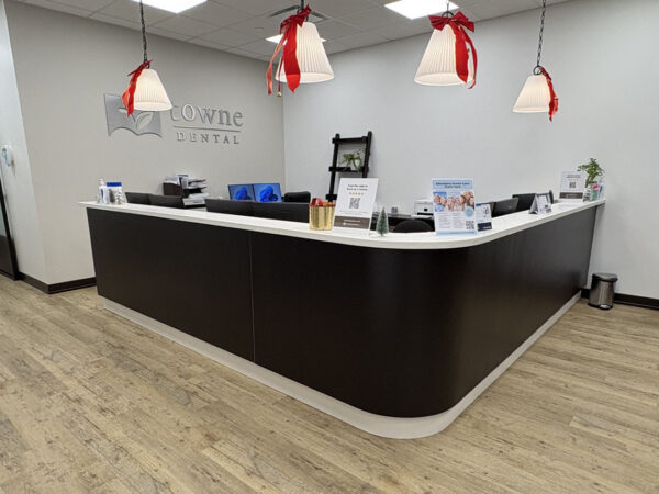 A modern dental clinic reception area with a sleek, dark counter. Three hanging lights with red bows provide a warm ambiance. Various informational brochures and decor items are arranged neatly on the countertop. The floor features light wood paneling.