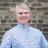 A smiling person with short gray hair wearing a light blue checkered shirt stands in front of a brick wall.