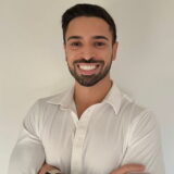 A person smiling warmly, wearing a white shirt, standing against a plain background with arms crossed.