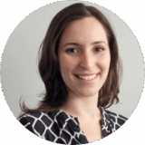 A smiling woman with long, dark hair is wearing a patterned blouse. She stands against a light wall, exuding warmth and approachability.