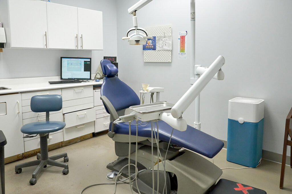 An interior view of a dental clinic featuring a blue dental chair, a treatment area with dental equipment, and a computer workstation. The space is clean and organized, designed for patient comfort and dental procedures.