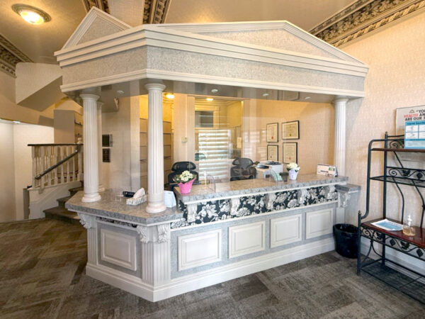 A dental reception area featuring a stylish front desk with columns, floral decor, and organized documents. The space is well-lit and has a welcoming atmosphere. Nearby is the cheerful 123Dentist Smiley, adding a friendly touch to the environment.