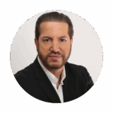 A professional man with a well-groomed beard and short hair is wearing a black suit jacket over a white shirt. He is looking directly at the camera with a serious expression against a plain background.