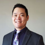 A professional man in a suit smiles warmly at the camera, exuding confidence and approachability. He has short black hair and is wearing a patterned tie with shades of purple. The background is neutral, enhancing his friendly demeanor.