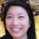 A person with long dark hair and sunglasses on their head is smiling warmly against a softly lit background.