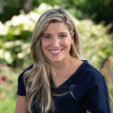 A person with long blond hair and a blue top smiling, sitting outdoors with greenery in the background.