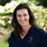 A smiling woman in a navy blue top stands outdoors, with greenery in the background. She appears friendly and professional, wearing attire that suggests a dental clinic setting.