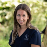 A smiling woman with long hair wears a dark blue dental uniform with a logo, set against a blurred green outdoor background.