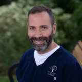 A man with a beard, wearing a navy dental uniform with the logo "Dentistry Dunbars," smiles warmly while sitting outdoors against a green leafy background.
