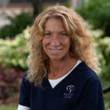 A smiling woman with curly hair wearing a navy blue dental uniform is sitting outdoors with green foliage in the background.