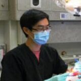 A dentist wearing a black uniform, surgical mask, and gloves is focused on a dental procedure in a clinic setting.