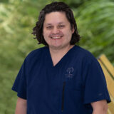 A person with dark hair in navy blue scrubs smiles outdoors with a blurred green background.