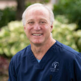 A smiling doctor in a blue dental uniform stands outdoors, with greenery blurred in the background.