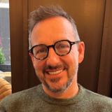 A person with glasses and a beard is smiling warmly, seated indoors against a dark background.