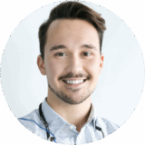 A friendly male dentist with a warm smile is wearing a light blue button-up shirt and a stethoscope around his neck. The background is neutral, emphasizing his approachable demeanor.