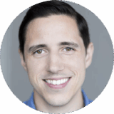 A smiling man with short dark hair and light skin is looking directly at the camera. He has a friendly expression and is dressed casually in a blue shirt. The background is soft and neutral, emphasizing his cheerful demeanor.