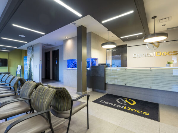 Modern dental clinic reception area with stylish seating, a sleek front desk, and ambient lighting. The room is bright and welcoming, featuring a large sign for "DentalDocs" and a flat-screen TV.