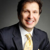 A smiling person in a dark suit and yellow tie against a neutral background.