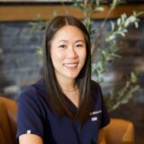 A woman in a navy blue uniform smiles warmly while sitting indoors, with a blurred plant and wall in the background.