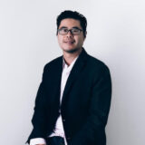 A man with glasses and a beard sits in a relaxed pose, wearing a dark blazer over a white shirt, against a plain white background.