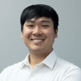 A smiling young man in a white shirt poses confidently against a light gray background, showcasing a friendly demeanor.