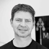 A smiling man with short dark hair is standing indoors, partially in front of a sign featuring a decorative lamp post design.