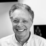 A smiling man with gray hair and a light-colored shirt looks directly at the camera. The background is blurred, and the image is in black and white.