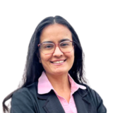 A person with long dark hair, glasses, and a warm smile, wearing a pink shirt and black blazer, stands against a plain white background.