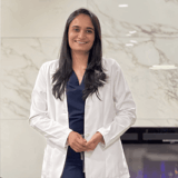 A professional smiling woman in a white lab coat stands confidently in a modern dental office. She has long dark hair and is positioned in front of a stylish marble wall, creating a welcoming atmosphere for patients.
