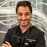 A smiling dentist in a black uniform, standing confidently with arms crossed, wearing dental loupes.