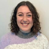 A person with curly hair smiles warmly, wearing a cozy sweater in shades of lavender and gray.