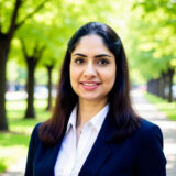 A woman in professional attire smiles while standing on a tree-lined path.