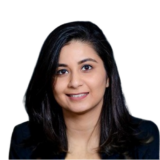 A person with long dark hair is smiling, wearing a dark jacket, against a plain background.