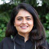 A woman with long dark hair and a warm smile, wearing a black shirt, stands outdoors against a backdrop of greenery.