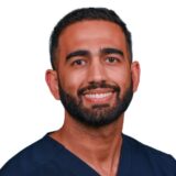 A man with a beard and short hair smiles while wearing a dark blue medical scrubs top. The background is plain white, enhancing his warm expression.