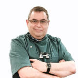 Un dentiste souriant, vêtu d'une blouse verte, se tient debout, les bras croisés, portant des lunettes et des instruments dentaires autour du cou. L'arrière-plan est d'un blanc uni, ce qui souligne son attitude amicale.