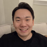 A person with short dark hair is sitting on a beige couch, smiling warmly at the camera.