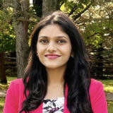 A woman with long black hair smiles confidently while wearing a pink blazer and a floral top. She is standing outdoors in a lush green setting with trees in the background.