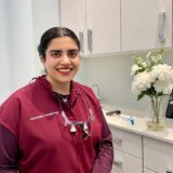 A smiling dental professional stands in a bright office surrounded by white flowers and modern dental equipment. She wears a maroon dental scrubs and glasses on her neck, exuding warmth and approachability.