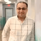 A smiling person stands casually in a brightly lit hallway, wearing glasses and a white checkered shirt. The background features soft green hues, creating a welcoming atmosphere.