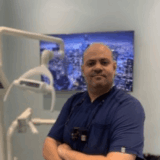 A dental professional stands confidently in a modern dental office, wearing blue scrubs and a dental loupes. Behind him is a large screen displaying a cityscape, adding a contemporary touch to the environment.