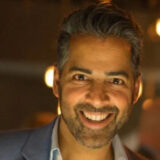 A man smiling warmly in a softly lit environment, wearing a dark suit with a light shirt.