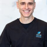 A smiling dentist in a black scrubs top stands against a light, marble background. His friendly expression conveys warmth and professionalism.