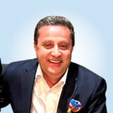 A smiling man in a dark suit and white shirt, with a pocket square, against a light blue background.