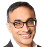 A person with glasses is smiling warmly, wearing a suit and a light shirt, set against a white background.